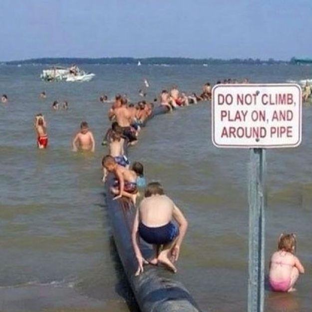 Funny warning and caution signs, warning sign next to large pipe in water stating it should not be climbed, beside a large crowd of people climbing the pipe and playing in the water
