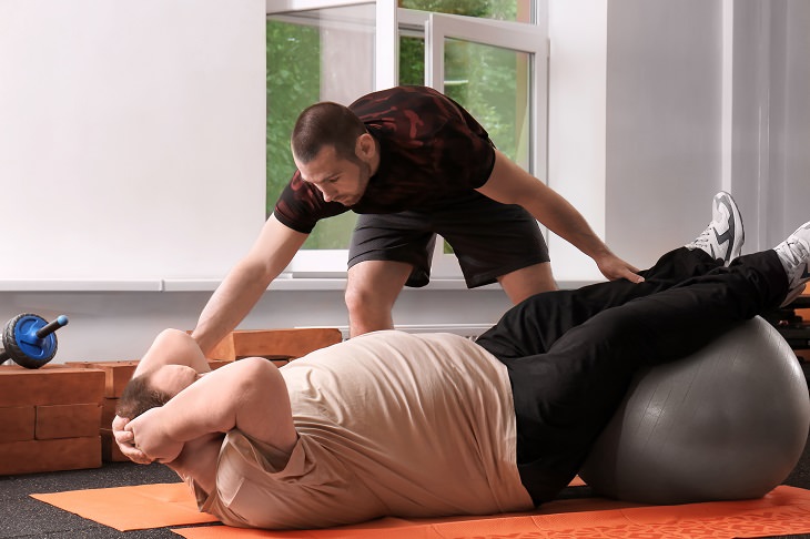 Exercising before food is beneficially to health. overweight man exercising in a gym with a trainer using a silver exercise ball