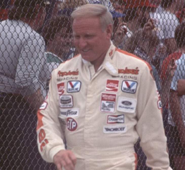 Top 19 NASCAR Race Drivers to Win Multiple Tracks, Cale Yarborough walking next to a fence in a white and red racing suit