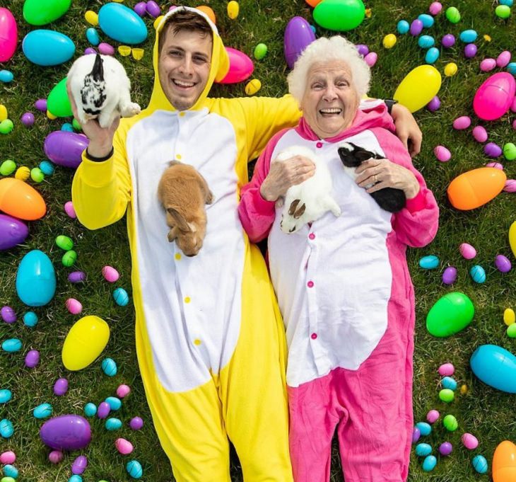 duo of grandmother and grandson, ross smith, wear fun costumes for social media, dressed in easter egg onsies costumes holding rabbits surrounded by easter eggs
