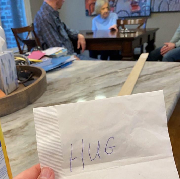 Funny and heartwarming pictures of birthday celebrations during the COVID-19 pandemic quarantine and lockdown, napkin with “HUG” written on it attached to a 6 foot long wooden stick, grandmother sitting 6 feet away at the table with other people