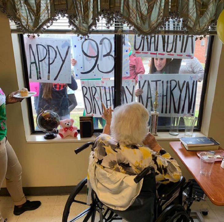 Funny and heartwarming pictures of birthday celebrations during the COVID-19 pandemic quarantine and lockdown, grandmother in wheel chair looking outside window at family holding signs that say “Happy 93rd Birthday Grandma Kathryn”
