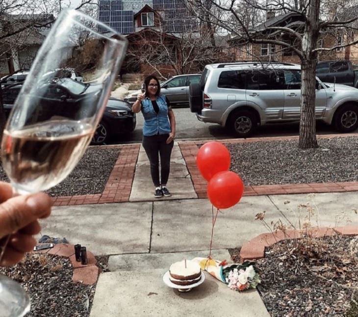 Funny and heartwarming pictures of birthday celebrations during the COVID-19 pandemic quarantine and lockdown, cake and flowers on ground in front of woman on sidewalk holding champagne glass