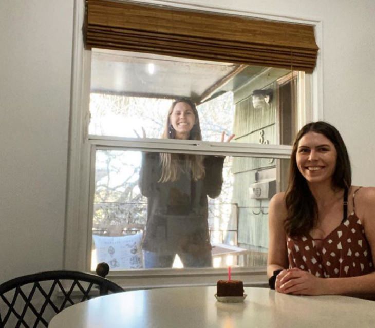 Funny and heartwarming pictures of birthday celebrations during the COVID-19 pandemic quarantine and lockdown, one sister stands outside window while the other sits inside with a piece of cake on the table