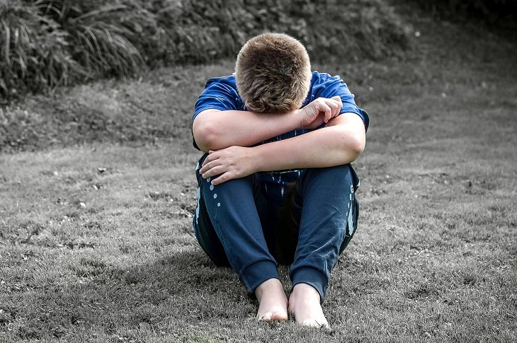 Debunking the myths about mental health, sad boy in blue t-shirt and blue pants sitting in the grass with face between hands and knees.