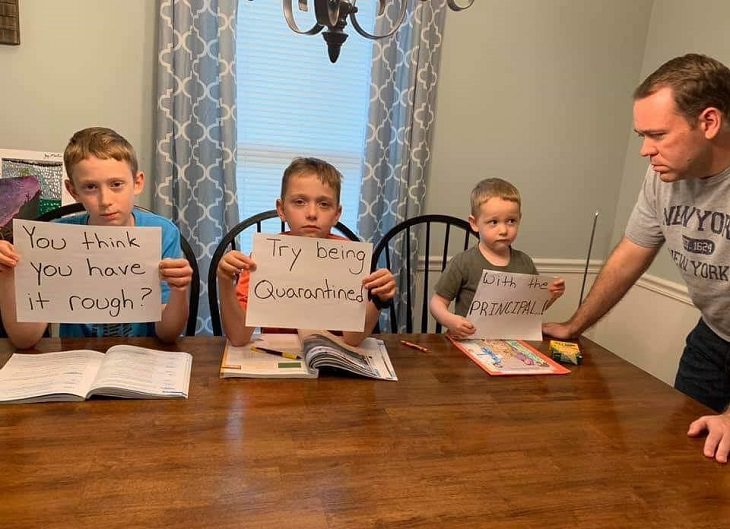 Funny signs related to the quarantine and lockdown caused by the COVID-19 (coronavirus) pandemic, Three young boys being watched by stern dad holding signs saying, “You think you have it rough? Try being quarantined with the principal.”