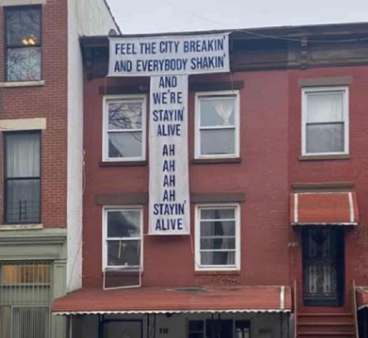 Funny signs related to the quarantine and lockdown caused by the COVID-19 (coronavirus) pandemic, banner hanging on building with altered lyrics for Stayin’ Alive by the BeeGees