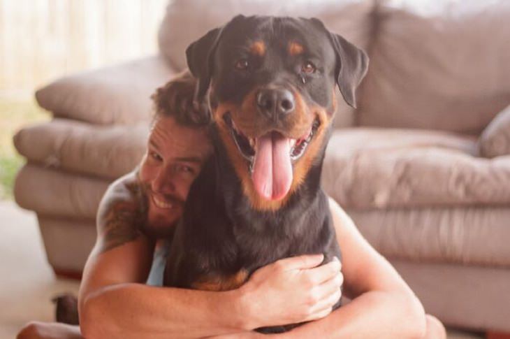 Photographs of smiling dogs, Black and brown rottweiler smiling at the camera while being hugged by owner