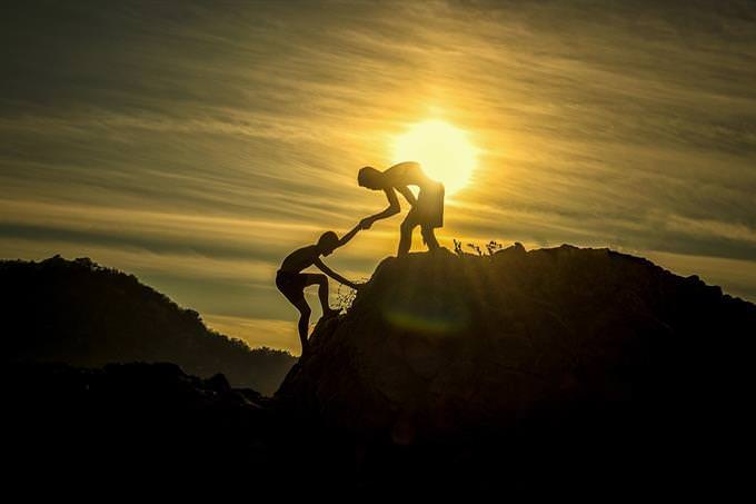 The silhouettes of a man helping his friend climb a mountain