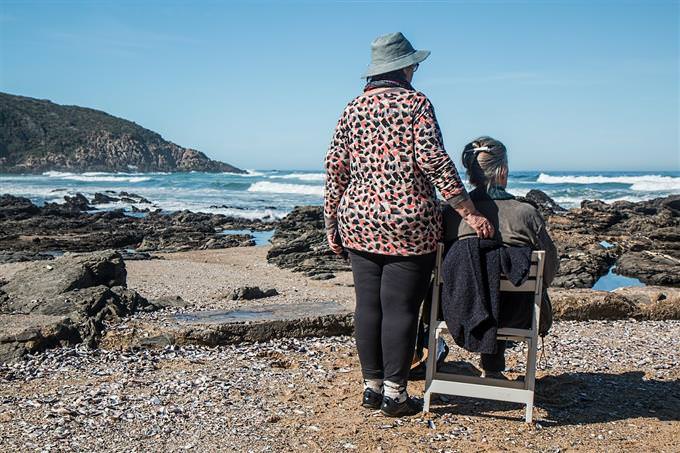 A woman standing next to a woman sitting on a chair at the beach