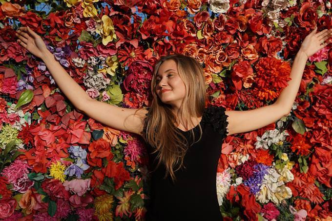 A woman with her arms spread sideways in front of a wall of flowers