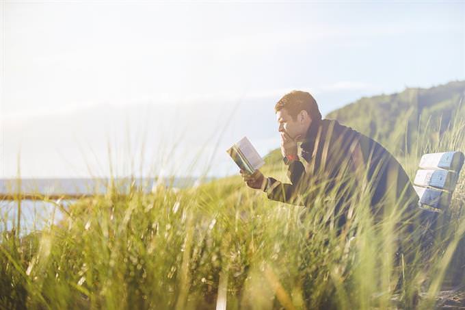 A man sits on a bench in a natural area and reads a book