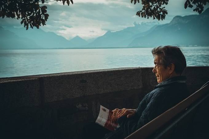 An elderly man sitting on the edge of a lake with a book