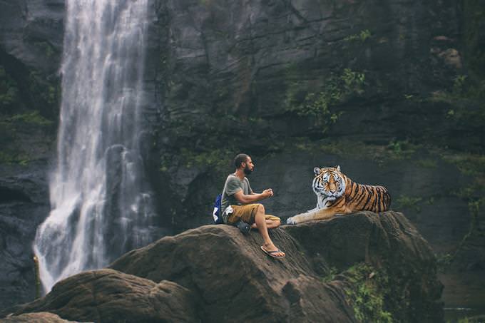 A man sitting beside a tiger in front of a waterfall