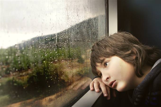 A boy leaning his head against a window while it’s raining outside