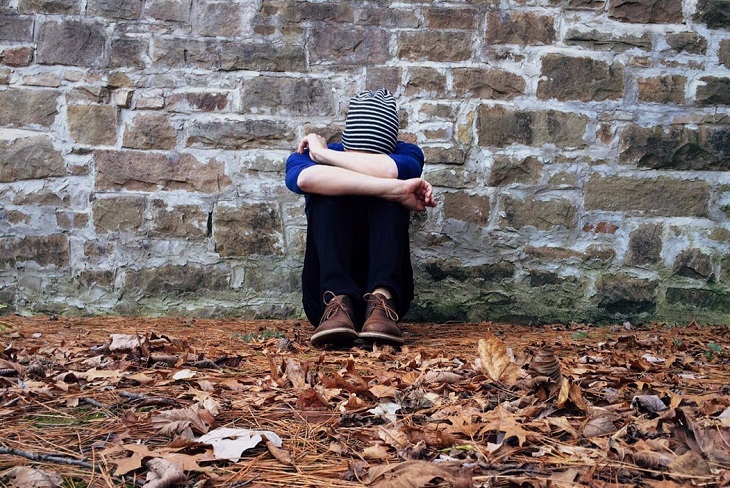 Fears of people-pleasers: a child sitting on a leaf-covered sidewalk, resting his head on his hands and leaning against a brick wall